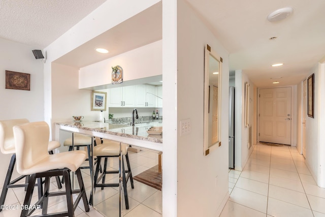 kitchen with light tile patterned floors, stainless steel fridge, a breakfast bar area, light stone countertops, and white cabinets