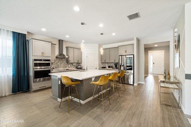 kitchen with a kitchen island with sink, stainless steel appliances, a kitchen bar, wall chimney exhaust hood, and light hardwood / wood-style flooring