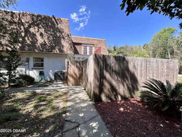 view of property exterior with central AC unit, fence, roof with shingles, mansard roof, and stucco siding