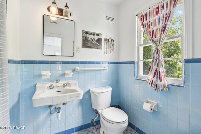 half bath with toilet, a sink, visible vents, tile walls, and wainscoting