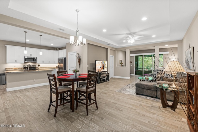 dining space with sink, a raised ceiling, light hardwood / wood-style floors, and ceiling fan with notable chandelier