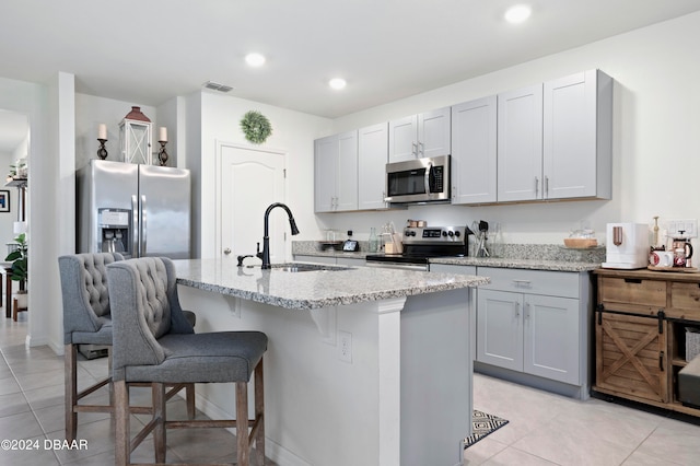 kitchen featuring stainless steel appliances, light tile patterned flooring, sink, light stone countertops, and an island with sink