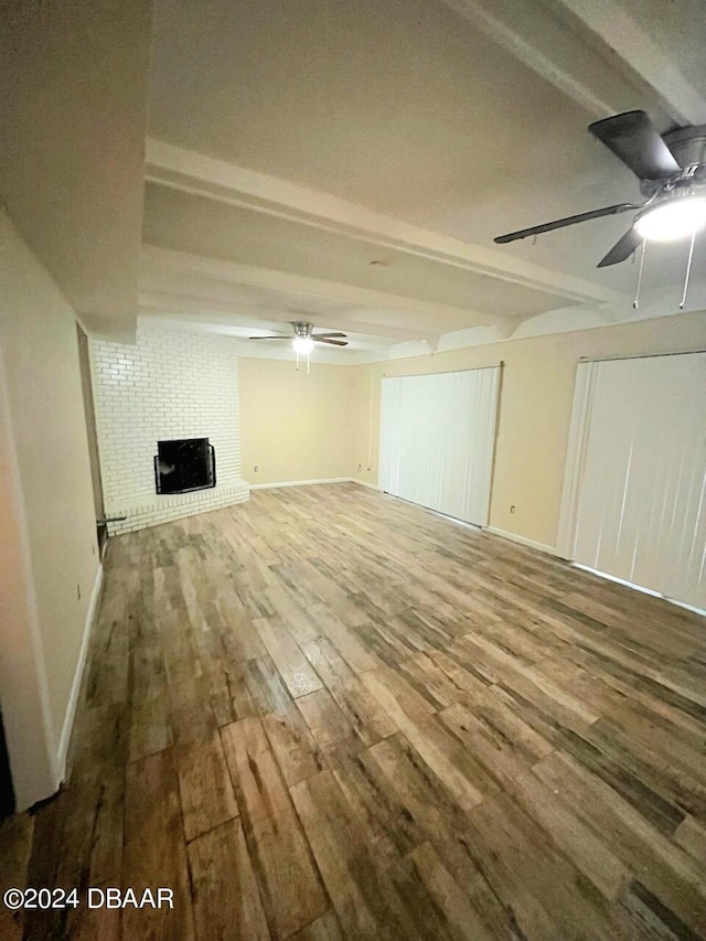 unfurnished living room featuring a brick fireplace, wood-type flooring, beamed ceiling, and ceiling fan