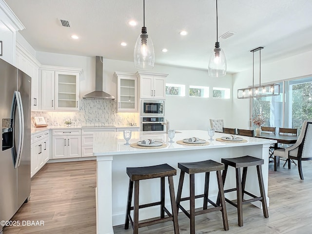 kitchen featuring wall chimney exhaust hood, hanging light fixtures, a center island with sink, stainless steel appliances, and white cabinets