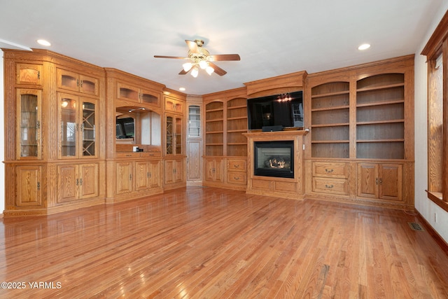 unfurnished living room featuring a glass covered fireplace, ceiling fan, light wood-style flooring, recessed lighting, and built in features