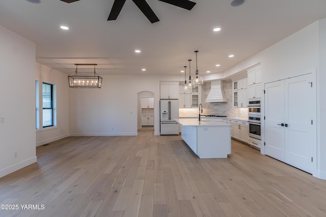 kitchen featuring high end white refrigerator, arched walkways, light countertops, glass insert cabinets, and a kitchen island with sink