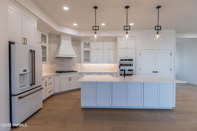 kitchen featuring white appliances, an island with sink, custom range hood, glass insert cabinets, and light countertops