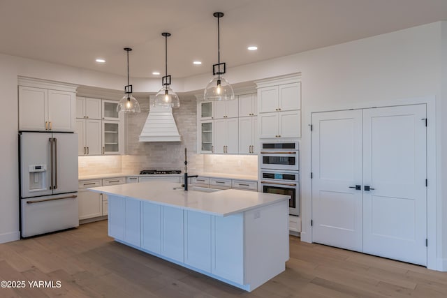 kitchen featuring decorative light fixtures, light countertops, glass insert cabinets, a kitchen island with sink, and white appliances