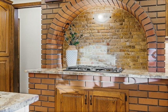 kitchen with brown cabinetry, stainless steel gas cooktop, light stone counters, and brick wall