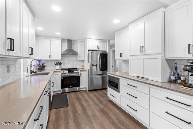 kitchen featuring wall chimney range hood, dark wood-style flooring, appliances with stainless steel finishes, white cabinets, and a sink