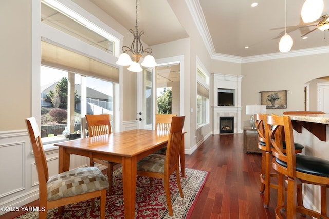 dining space featuring ornamental molding, dark wood finished floors, ceiling fan with notable chandelier, a wainscoted wall, and a tile fireplace