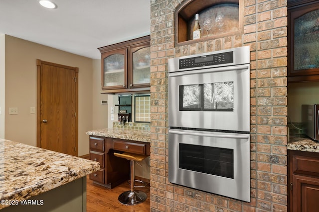 kitchen featuring stainless steel double oven, glass insert cabinets, light stone countertops, and wood finished floors