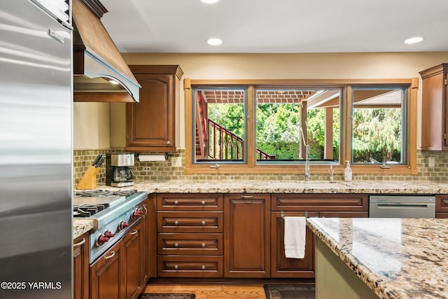 kitchen featuring premium range hood, light stone counters, a sink, stainless steel appliances, and decorative backsplash