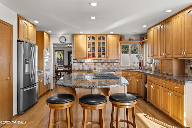 kitchen with appliances with stainless steel finishes, glass insert cabinets, a sink, dark stone counters, and a kitchen bar