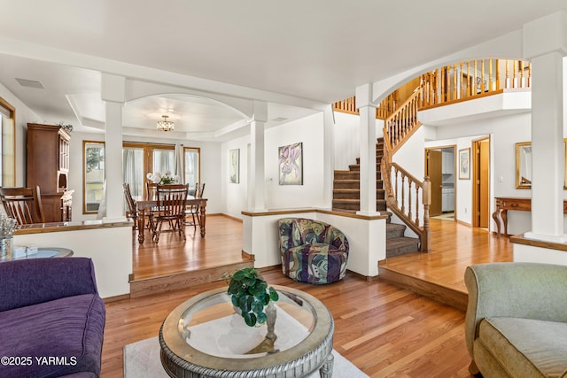 living area with light wood-type flooring, decorative columns, a tray ceiling, and stairs