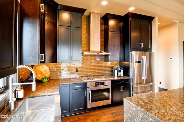 kitchen featuring light stone counters, stainless steel appliances, tasteful backsplash, a sink, and wall chimney exhaust hood