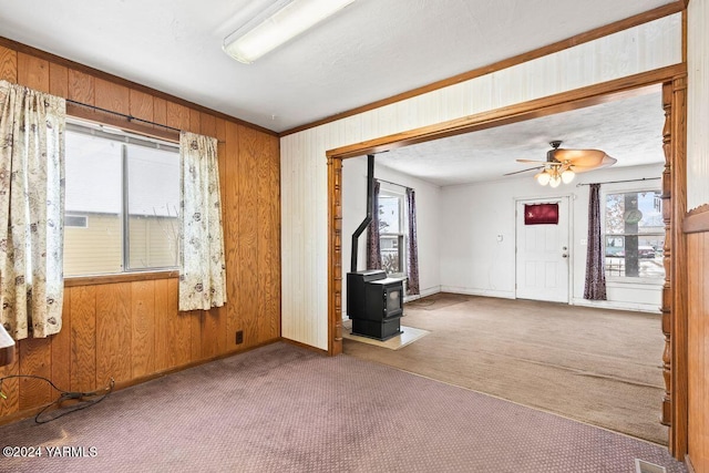 empty room featuring a wood stove, ceiling fan, wooden walls, and light colored carpet