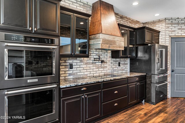 kitchen with light stone counters, stainless steel appliances, custom exhaust hood, dark wood-style floors, and glass insert cabinets