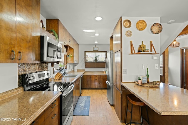 kitchen featuring light wood finished floors, a peninsula, a sink, stainless steel appliances, and brown cabinets