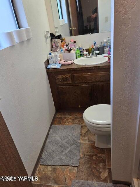 half bathroom featuring toilet, a textured wall, vanity, and stone tile flooring