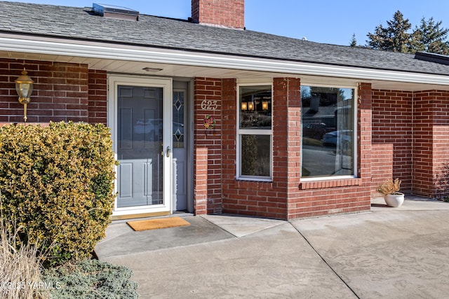 doorway to property with a shingled roof, a chimney, and brick siding