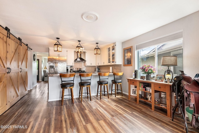 kitchen featuring a barn door, wall chimney exhaust hood, white cabinetry, a peninsula, and stainless steel refrigerator with ice dispenser