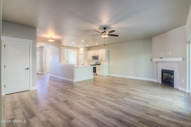 unfurnished living room featuring light wood finished floors, arched walkways, baseboards, ceiling fan, and a fireplace