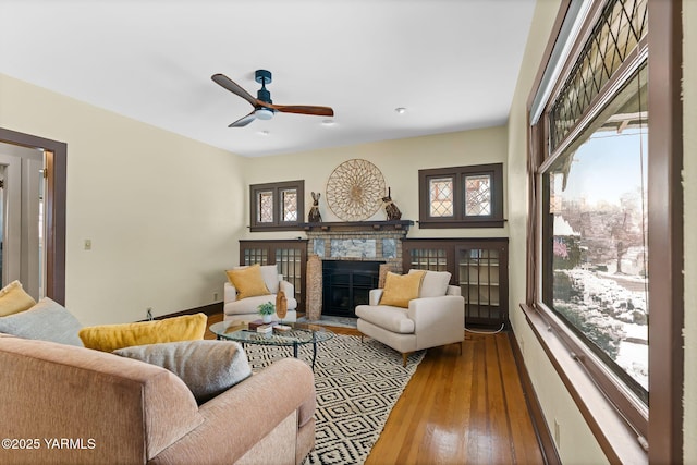 living area featuring baseboards, a fireplace with flush hearth, a ceiling fan, and wood finished floors