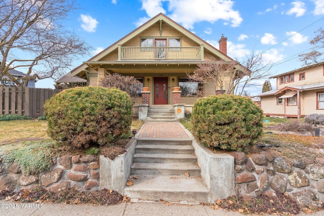 view of front of house with covered porch, a chimney, a balcony, and fence