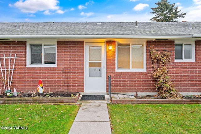 doorway to property featuring brick siding, a lawn, and roof with shingles