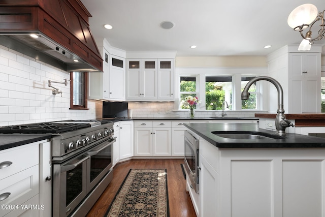 kitchen featuring a center island with sink, custom exhaust hood, stainless steel appliances, dark countertops, and glass insert cabinets