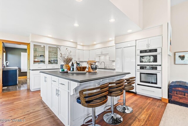 kitchen with dark countertops, a kitchen breakfast bar, white cabinetry, white double oven, and decorative backsplash