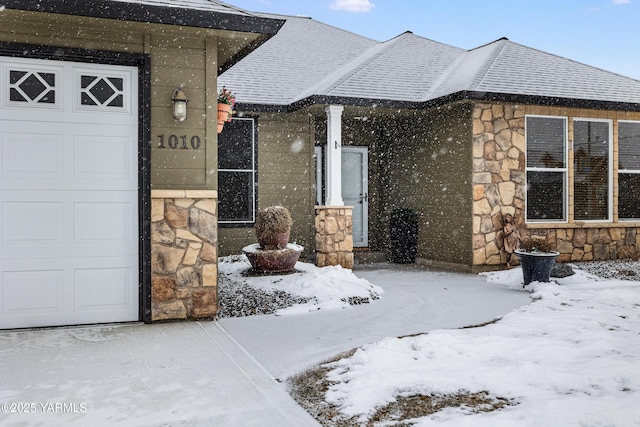 snow covered property entrance with a garage, stone siding, and roof with shingles