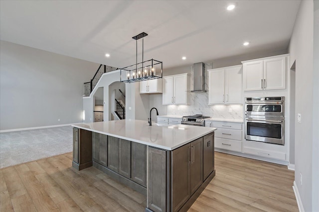 kitchen featuring wall chimney exhaust hood, white cabinetry, an island with sink, and stainless steel appliances