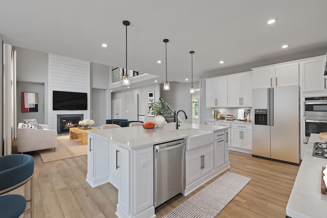 kitchen featuring a center island with sink, decorative light fixtures, a large fireplace, white cabinetry, and stainless steel appliances