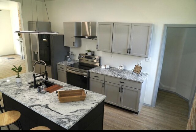 kitchen with stainless steel appliances, gray cabinets, light wood-type flooring, an island with sink, and wall chimney exhaust hood