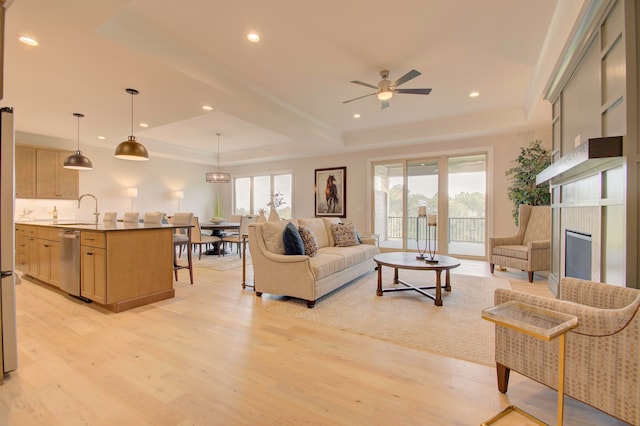 living room with ceiling fan, a raised ceiling, sink, a tile fireplace, and light wood-type flooring