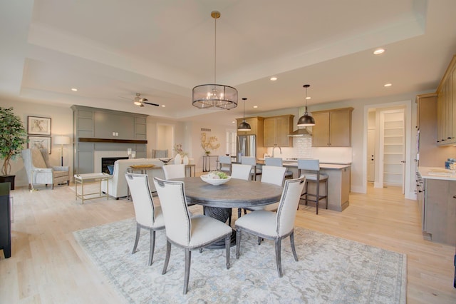 dining area with ceiling fan with notable chandelier, a tiled fireplace, a tray ceiling, light hardwood / wood-style flooring, and sink