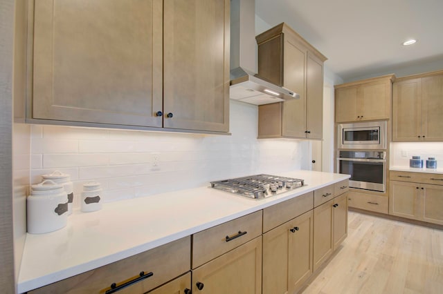 kitchen with appliances with stainless steel finishes, tasteful backsplash, wall chimney exhaust hood, light brown cabinets, and light hardwood / wood-style flooring