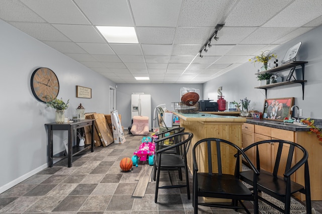 bar featuring a sink, white fridge with ice dispenser, wet bar, a drop ceiling, and baseboards