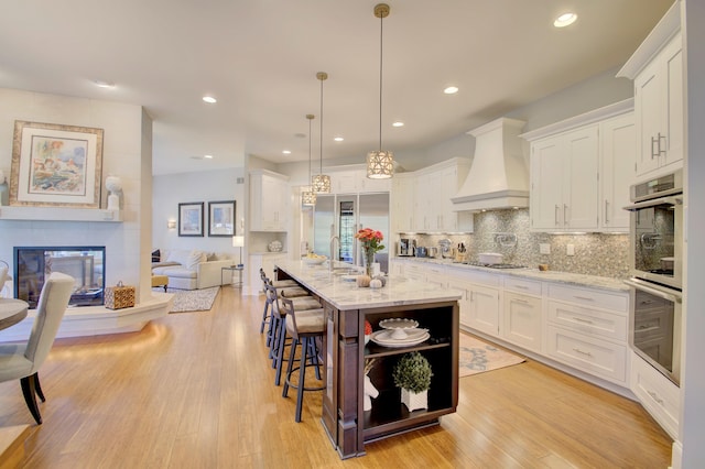 kitchen featuring an island with sink, pendant lighting, white cabinetry, and custom range hood