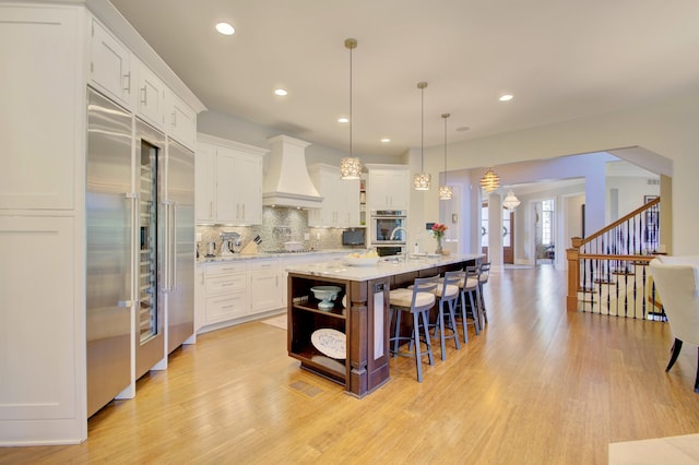 kitchen with custom exhaust hood, hanging light fixtures, a kitchen island with sink, a breakfast bar area, and white cabinets