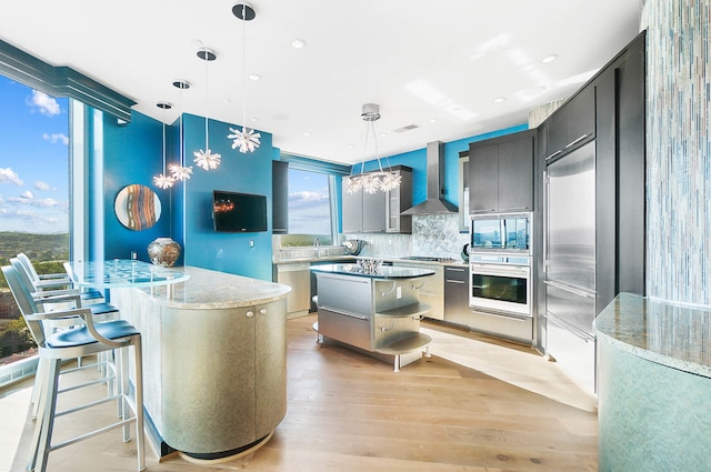 kitchen featuring wall chimney exhaust hood, hanging light fixtures, a center island, appliances with stainless steel finishes, and light hardwood / wood-style floors