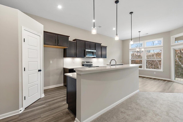 kitchen with decorative light fixtures, light countertops, appliances with stainless steel finishes, a kitchen island with sink, and a sink