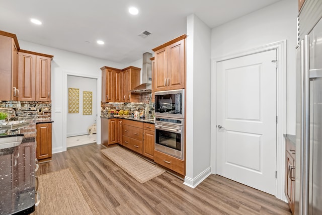 kitchen featuring wall chimney range hood, tasteful backsplash, dark stone counters, appliances with stainless steel finishes, and hardwood / wood-style flooring