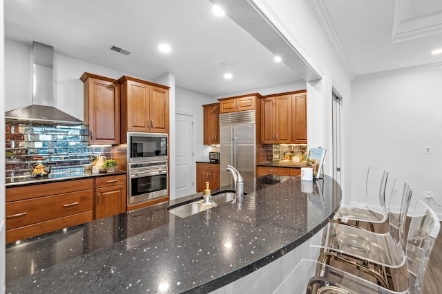 kitchen with sink, wall chimney exhaust hood, built in appliances, dark stone countertops, and decorative backsplash