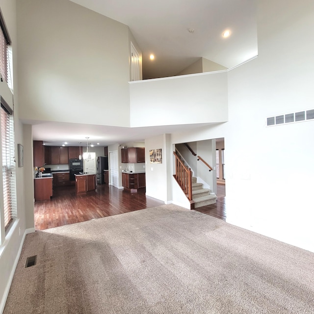 unfurnished living room featuring dark colored carpet, stairway, a towering ceiling, and visible vents