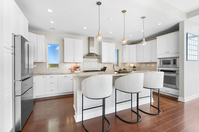 kitchen featuring a center island, white cabinetry, wall chimney range hood, stainless steel appliances, and hanging light fixtures