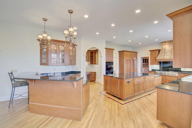 kitchen with custom exhaust hood, a kitchen breakfast bar, dark stone countertops, and light wood finished floors