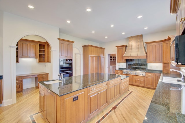 kitchen with a sink, custom exhaust hood, a kitchen island with sink, and dobule oven black